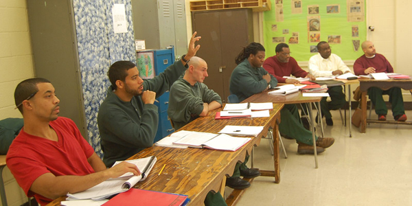 seven of the students from the prison seated at a long desk, one of them is raising his hand