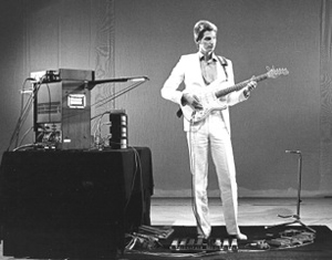 Dresher in white suit playing electric guitar surrounded by electronic equipment.