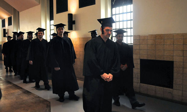 A group of inmates wearing graduation caps and gowns walks down a prison corridor.