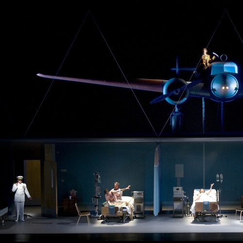 A staged scene from Amelia, in the center are two hospital beds, to the far left a man in uniform walks through a door and above it all is an old propeller airplane and its pilot.