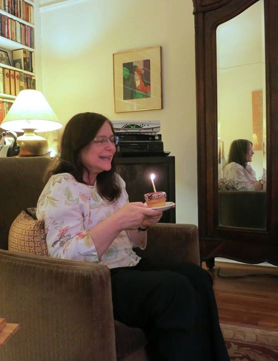 Deborah Atherton seated and holding a piece of brthday cake with a lit candle on top.