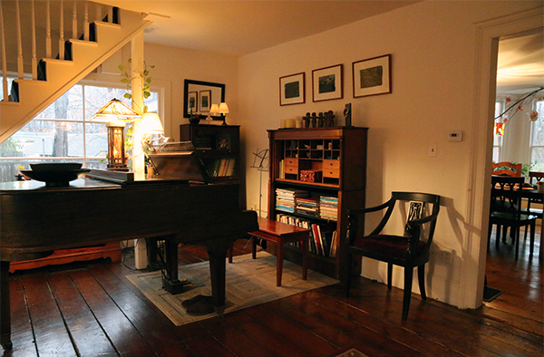 Photo of interior of room with grand piano, shelves, chair, and wooden floor.
