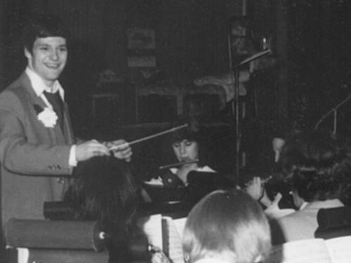 Historic photo of a teenaged Daron Hagen holding a baton as young musicians play instruments. 