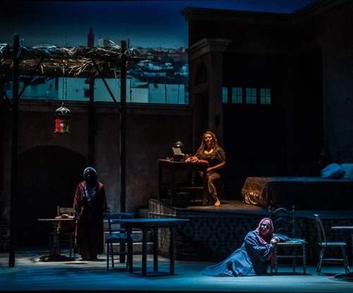 A darkly lit stage with trhee women: one standing, one sitting at a desk, and another on the floor leaning against a chair.