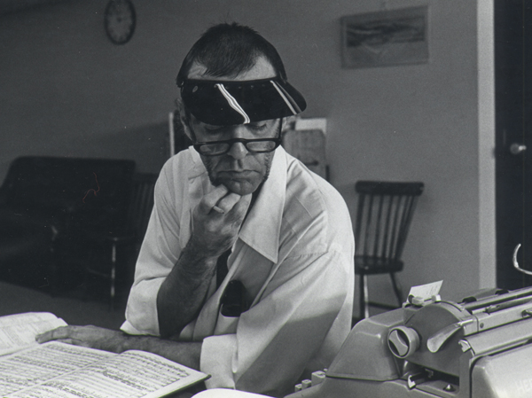 Ezra Sims at work composing wearing a dealer shade, seated at a desk with a manuscript score.