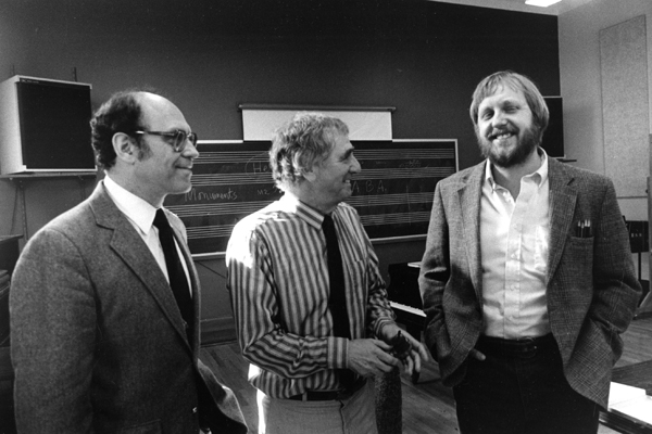 Adler, Rands and Schwantner standing and talking in a classroom in front of a blackboard with musical staves across it.