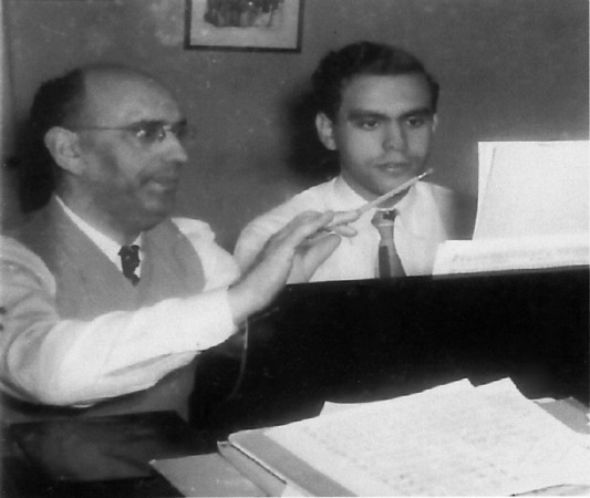 A young Samuel Adler sitting at a piano looking at sheet music while his father Hugo sitting next to him is speaking and has his hands raised as if conducting.