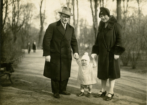 Sam Adler as a young child walking in a park with his father holding his right hand and his mother holding his left hand.