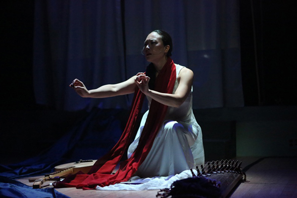 Jen Shyu, wearing a traditional white Asian dress and a red scarf sitting on the stage surrounded by a moon lute and a zither singing and making hand gestures