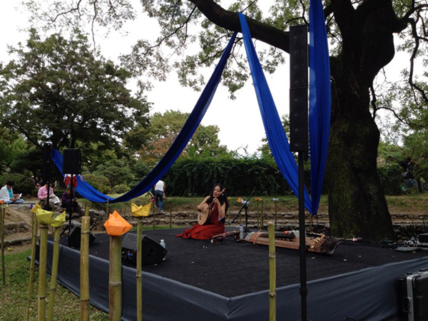 Jen Shyu singing and playing a moon lute on an outdoor stage in a park.