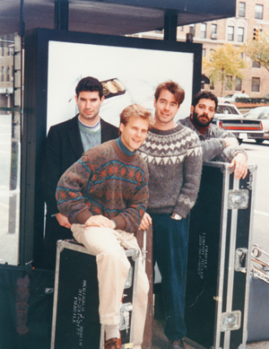 Historic photo of the four original members of PRISM standing in front of a bus kiosk with instrument cases.