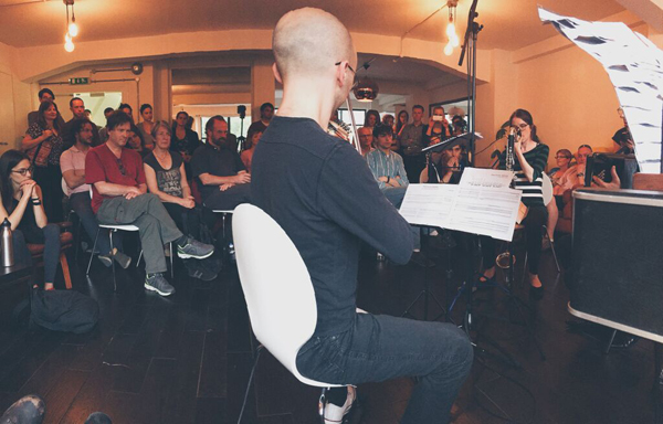 Photo of a man sitting and playing a violin as audience members seated around the room listen.