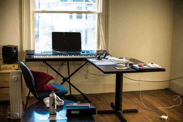 Photo of a songwriter's workspace. A digital keyboard synthesizer on a tripod positioned by a window perpendicular to a desk; on the desk is a notebook, laptop, headphones, and glass of water.