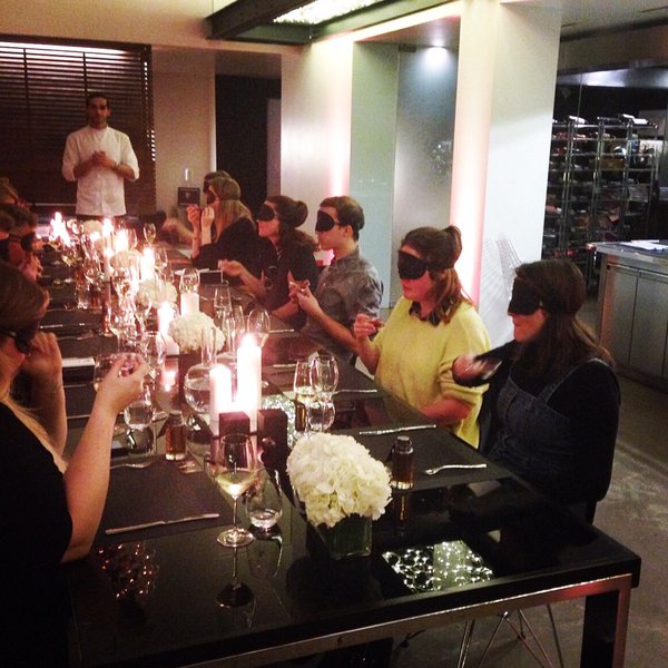 Blindfolded patrons sit around a table and eat various food items, smell perfume, and listen to music.