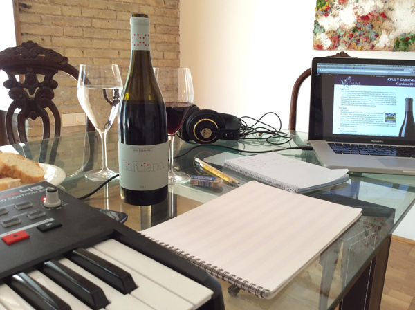 A bottle of wine and a wine glass on a tale alongside an electronic keyboard, music notation paper, headphones, and a computer screen.