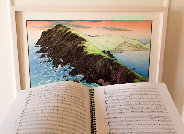 A musical score in front of a framed artwork showing a landscape scene of a mountain surrounded by water.