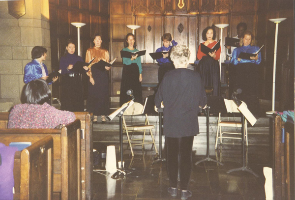 A conductor and ight sopranos singing in front of music stands at a church.