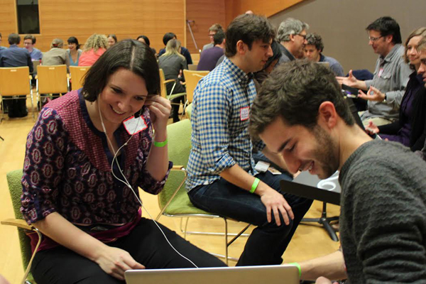 People sitting across from each other in pairs in a room. In front a woman listening to music with an earbud that is emanating from a man's laptop.