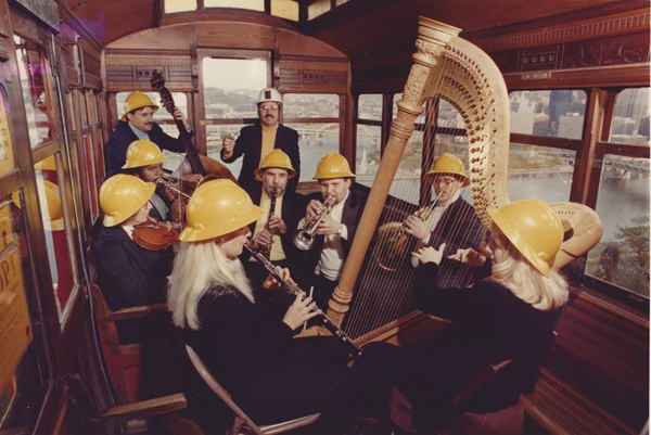 A group of eight musicians wearing yellow hard hats and playing various instruments (doublebass, harp, clarinet, trumpet, French horn, violin) conducted by a young David Stock wearing a white hard hat in what looks like a cable car.