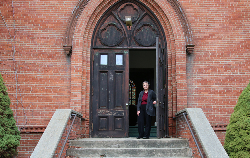 Mary Jane Leach standing outside the entrance of the church she lives in; a stained glass window is visible through the door.