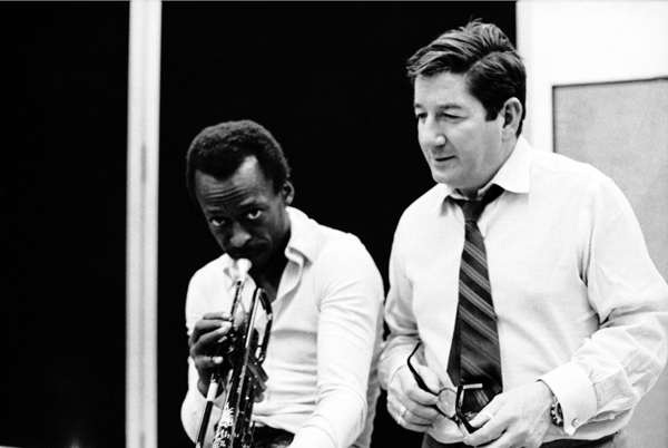 Miles Davis and Teo Macero standing together each wearing a white shirt and a tie; Miles is also playing trumpet