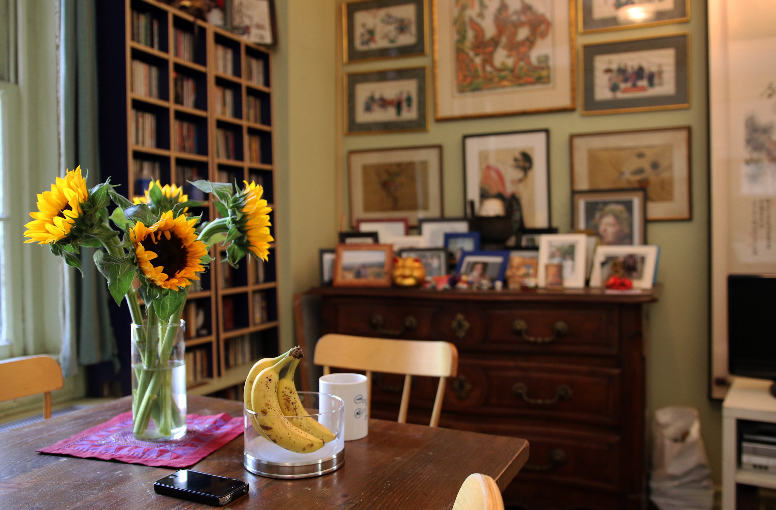 John King's kitchen. A table with flowers and some bananas, a bookcase, and a wall filled with framed photographs.