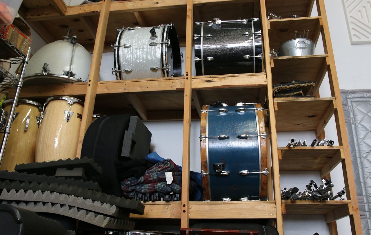 shelves of drums containing a timpano (upside down), three side drums, a pair of congas, and various drum stands.