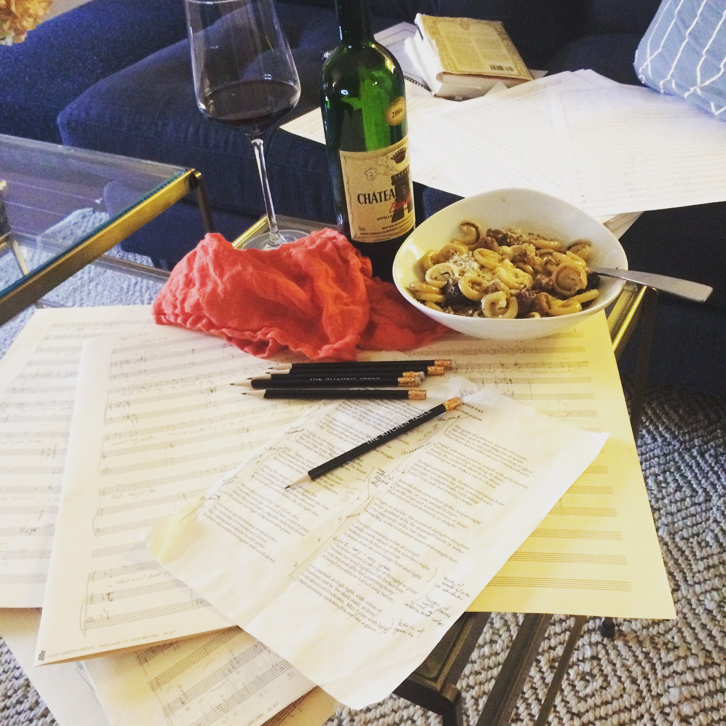 A table with pages of score manuscript paper, pens, a glass of red wine, a wine bottle, and a bowl of pasta