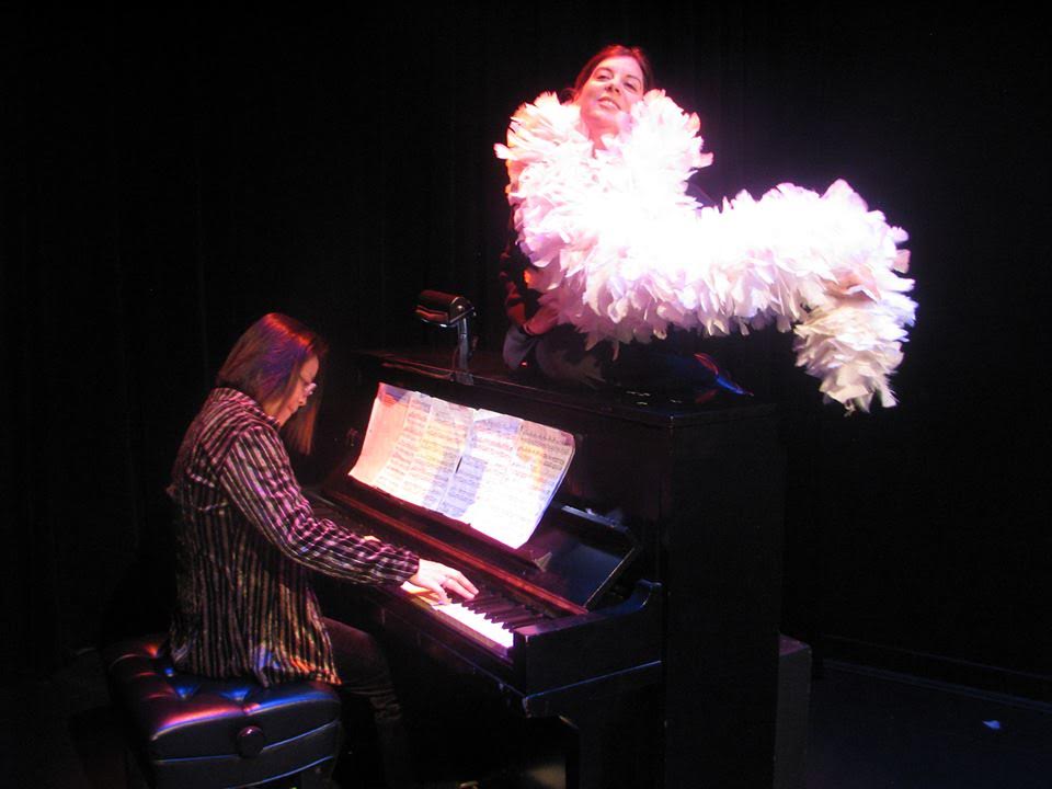Missy Fogarty in a boa sitting on top of a piano singing and accompanied at the piano by Shoko Nagai.