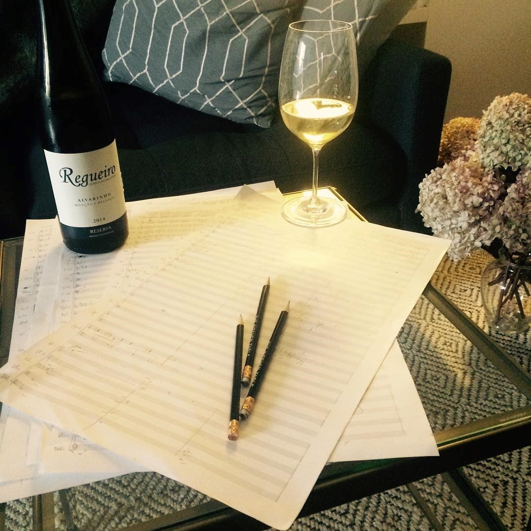 A wine bottle and a glass filled with white wine next to some page of music score paper and a few pencils.