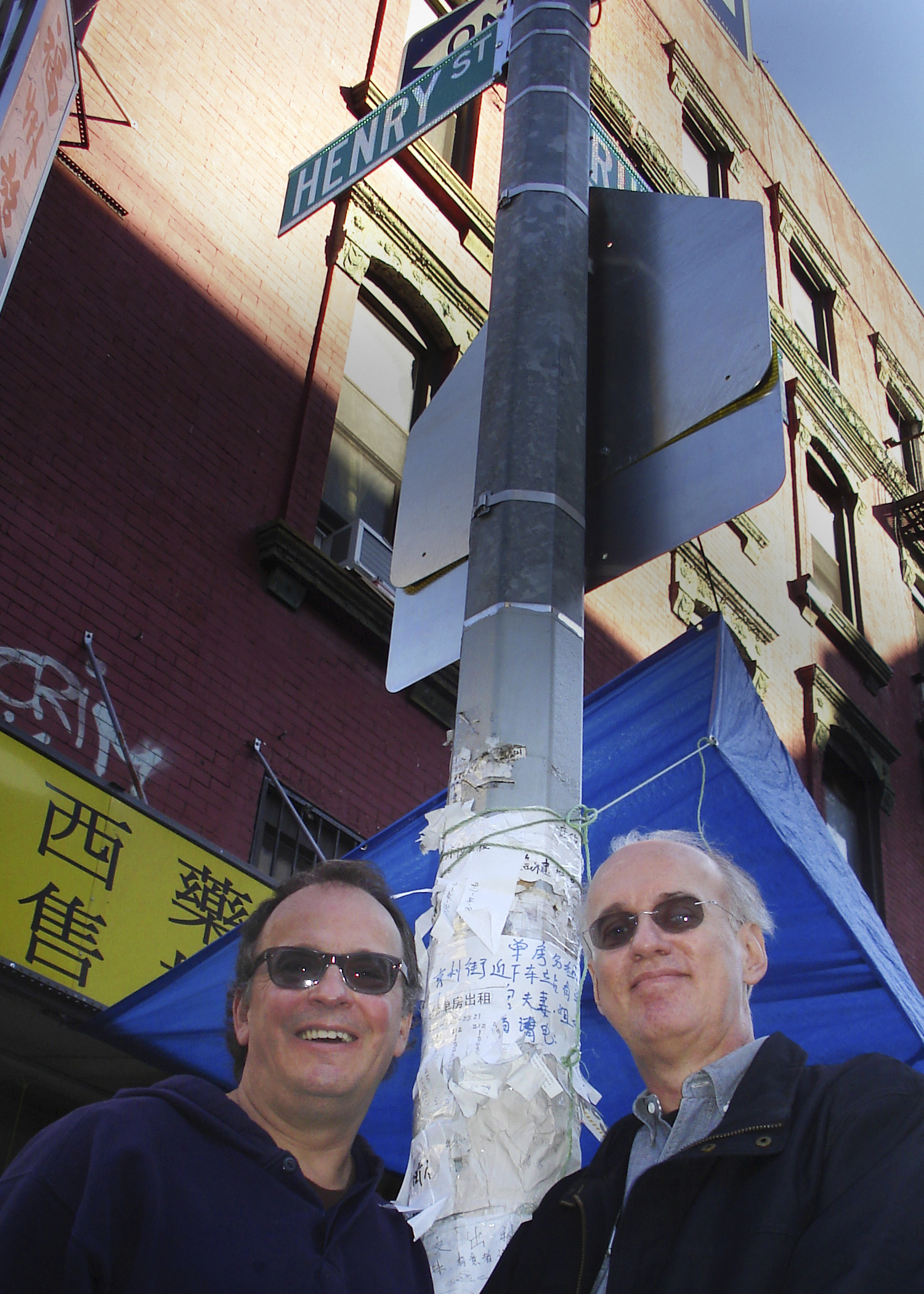 Phil Kline and Tom Steenland both wearing sunglasses and standing on opposite sides of a traffic pole on a city street corner.