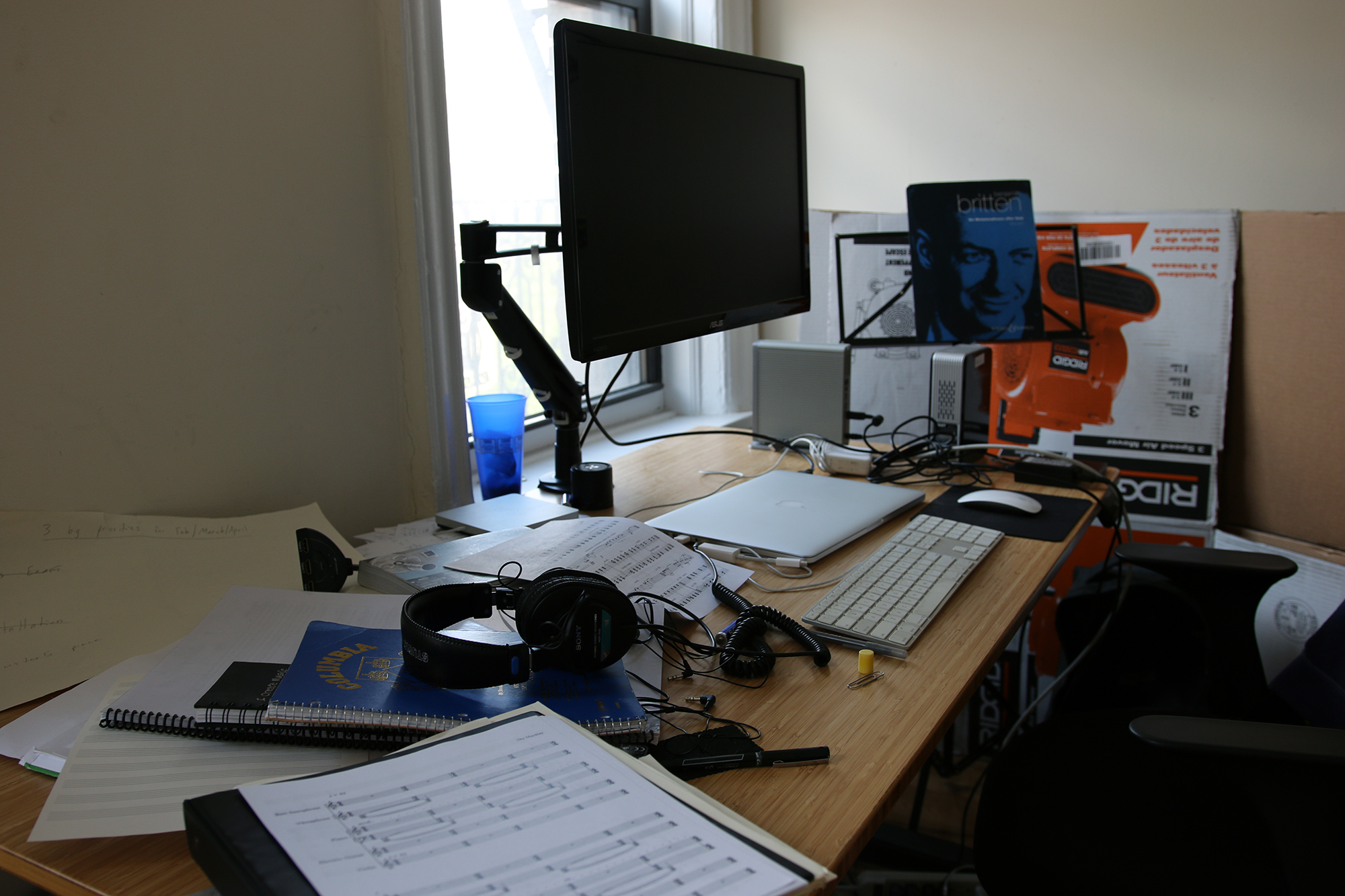 A work table with a closed laptop, an additional computer keyboard and large-scale monitor, headphones, and printed musical scores.