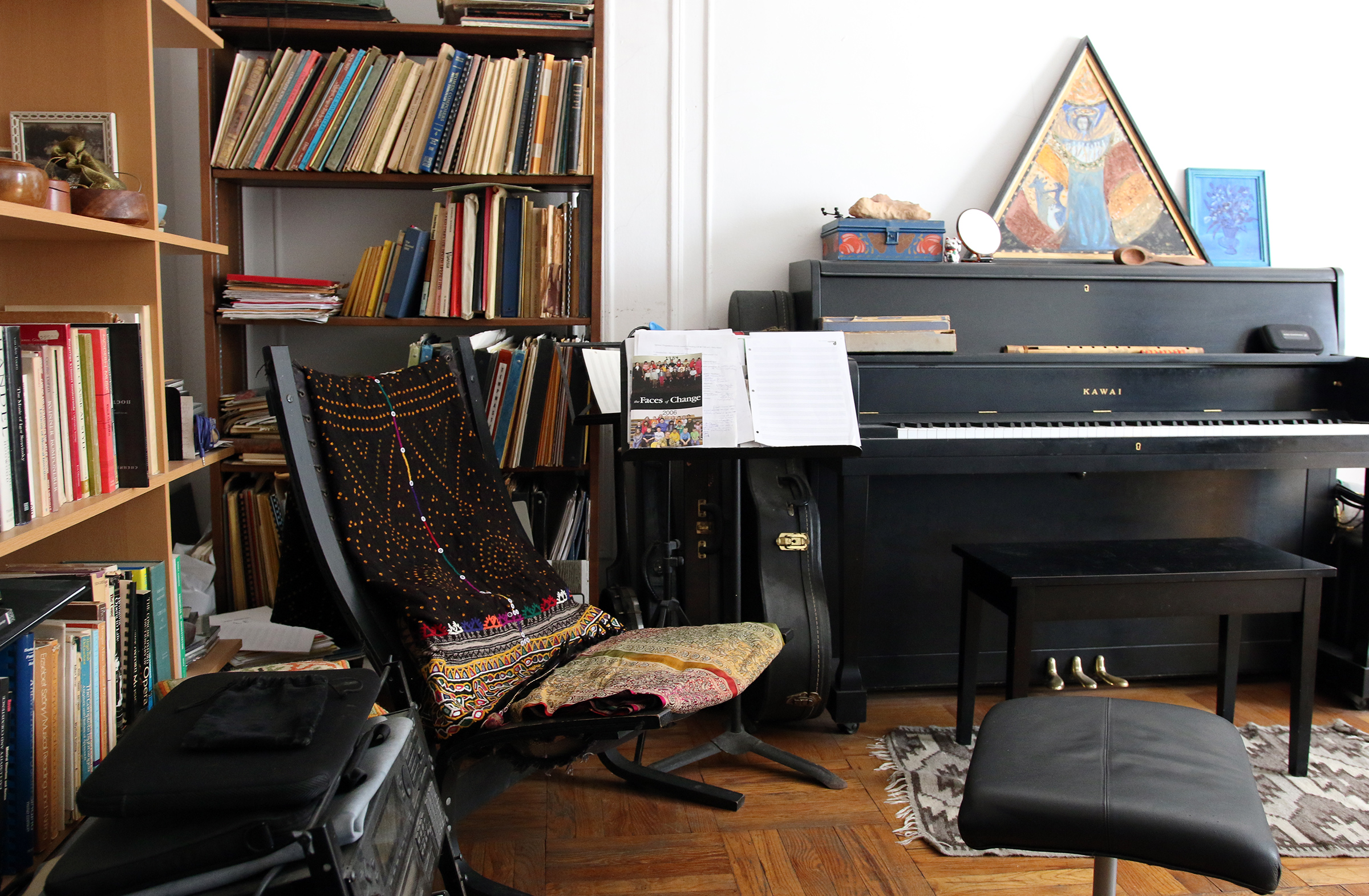 A room with shelves of books, a chair, and in the middle, an upright piano with a triangular painting on top of it.