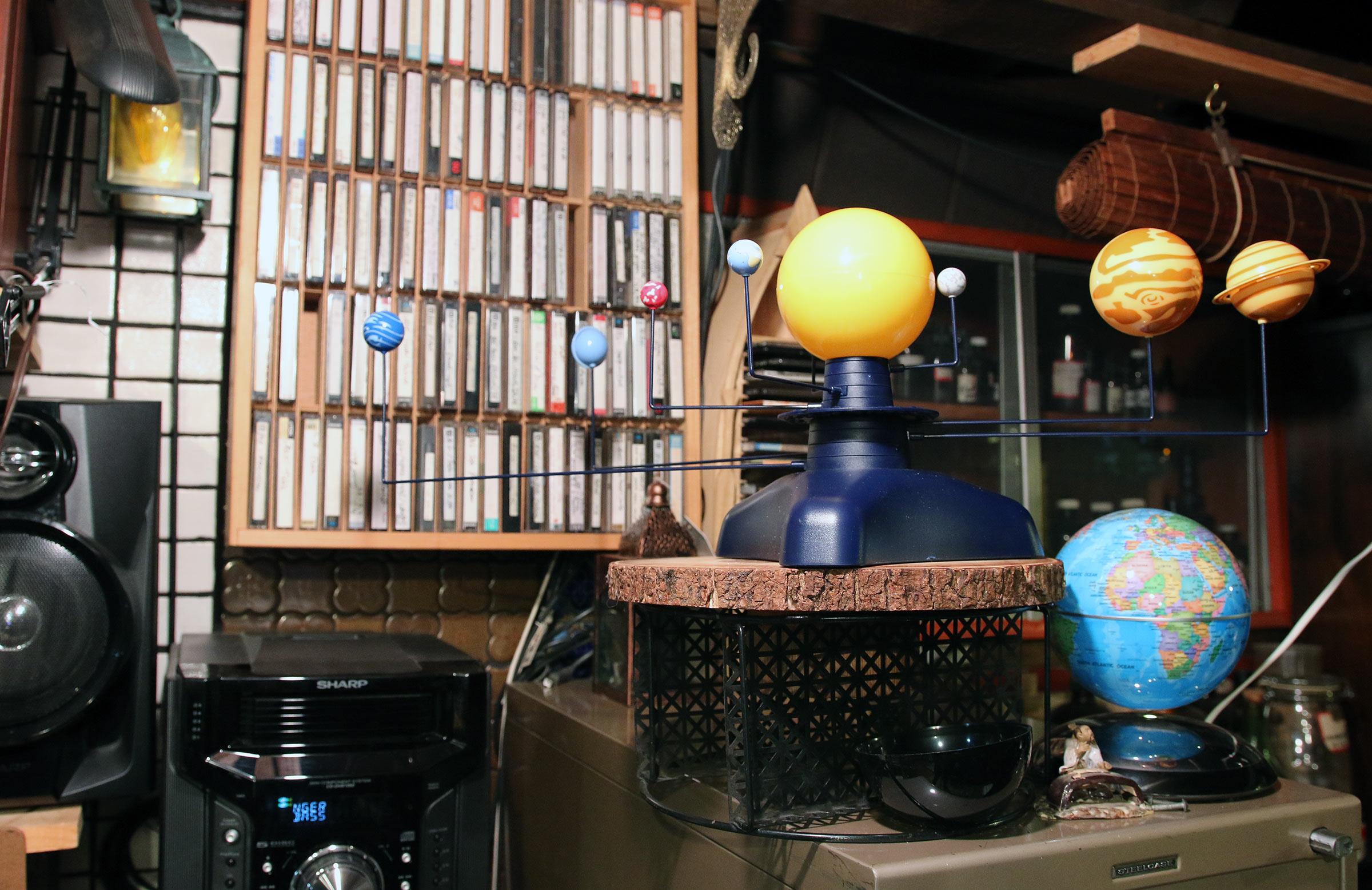 A globe of the earth as well as globes of other planets and satellites in our solar system sits in front of a shelf filled with cassettes of Milford Graves's performances.
