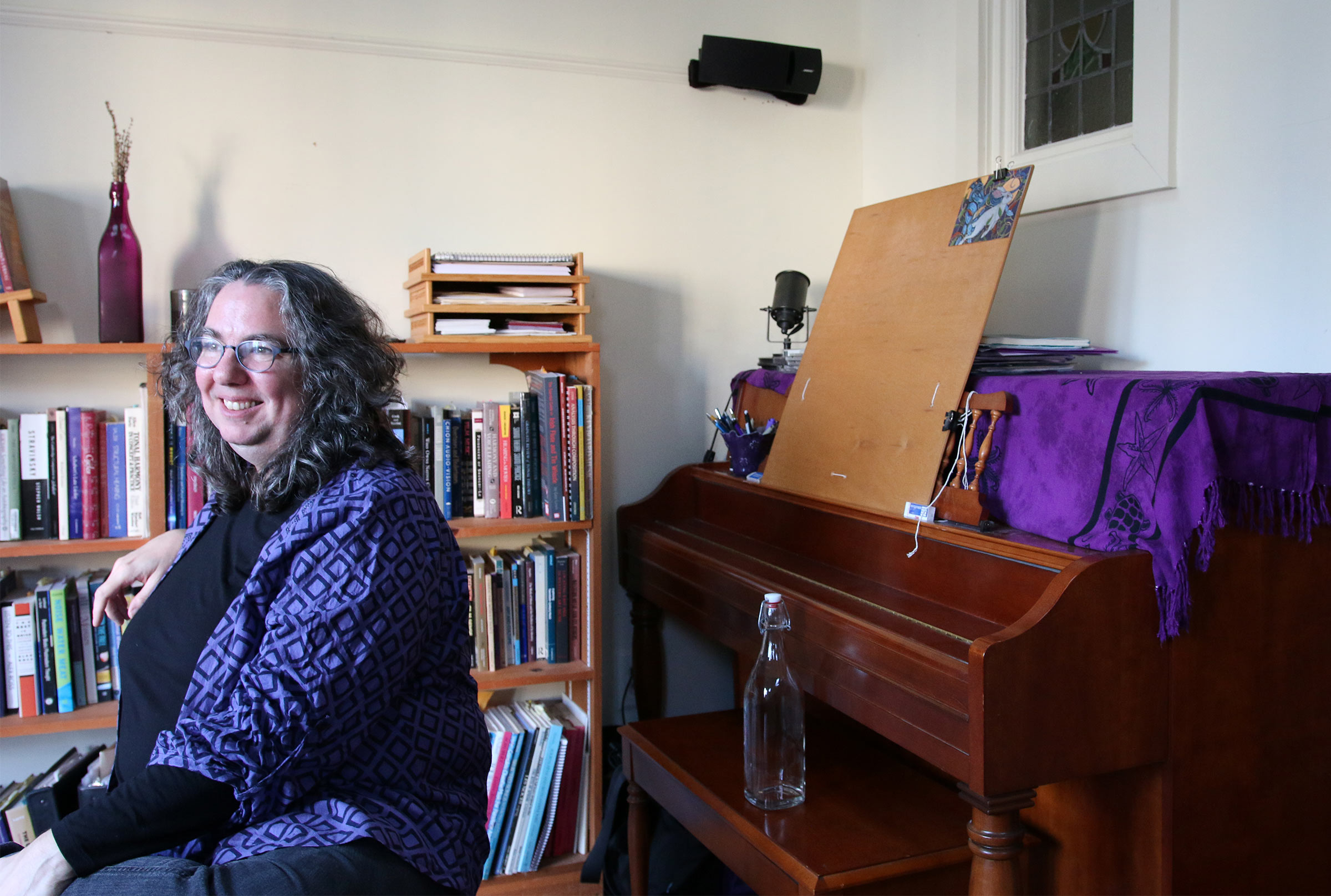 Barbara White sitting in her studio with her upright piano in back of her.