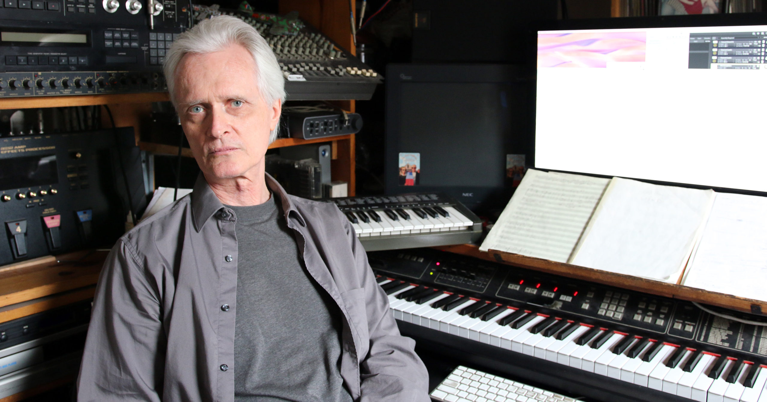 Scott Johnson sitting at his compositional work station surrounded by electric keyboard, mixers, music notation paper, and a large computer screen.
