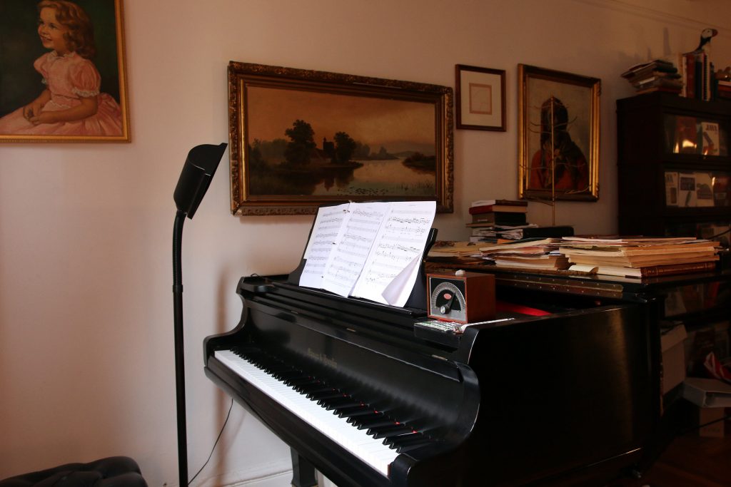 In front of a group of paintings there is a grand piano in Beth Anderson's living room with piles of sheet music on the lid and stand.