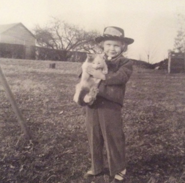 Beth Anderson, as a young girl, holding a cat and hearing a hat outside on a farm.