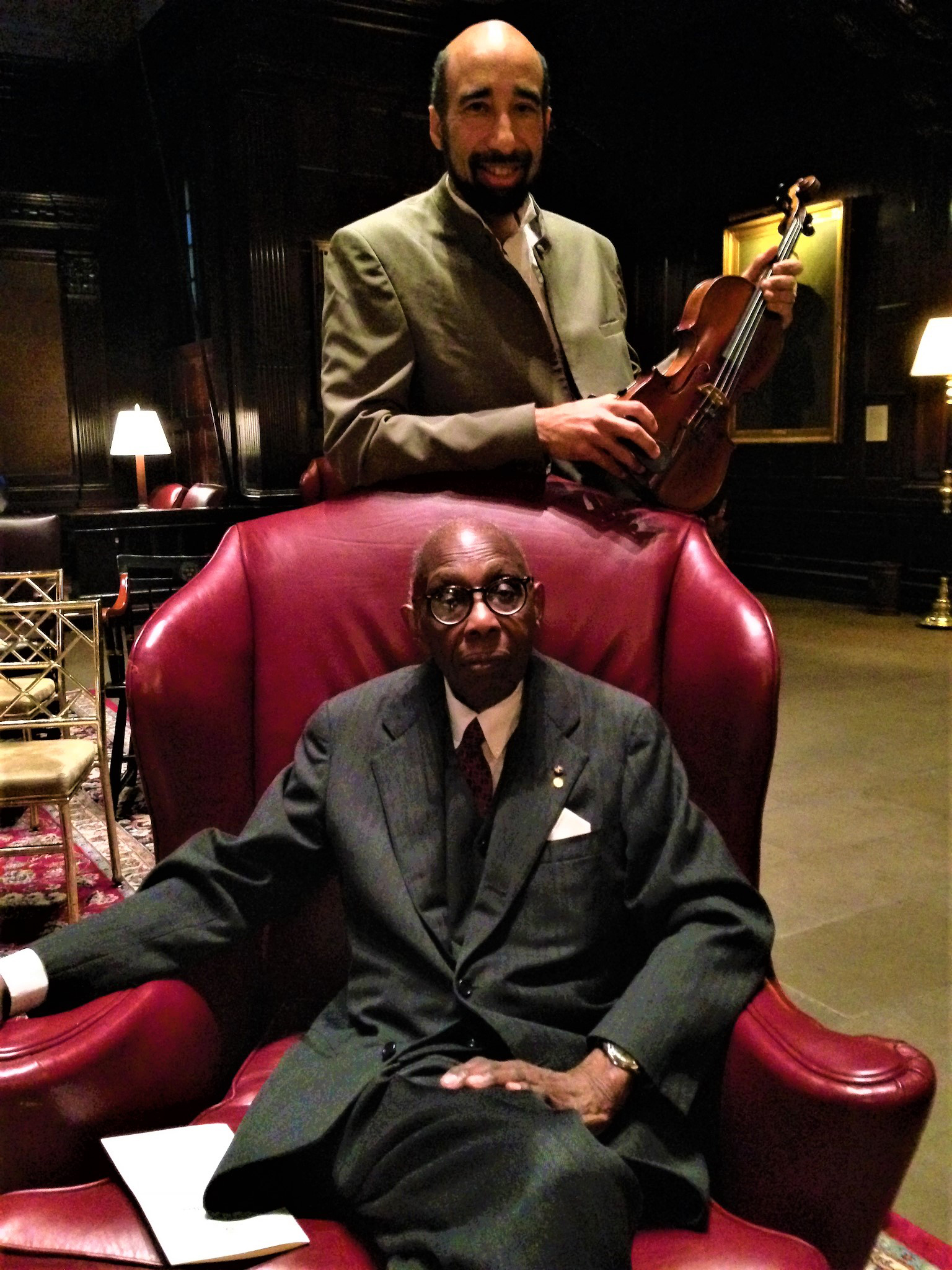 Gregory T. S. Walker standing and holding a violin with George Walker seated on a red chair.