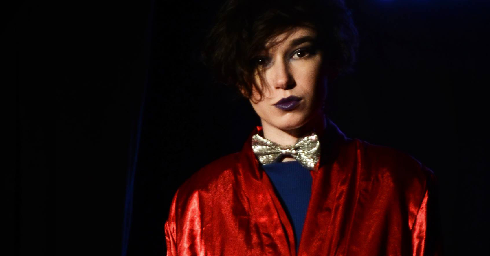 A woman in a dark red coat, blue shirt and dark lipstick posing as the role of a prince for an opera