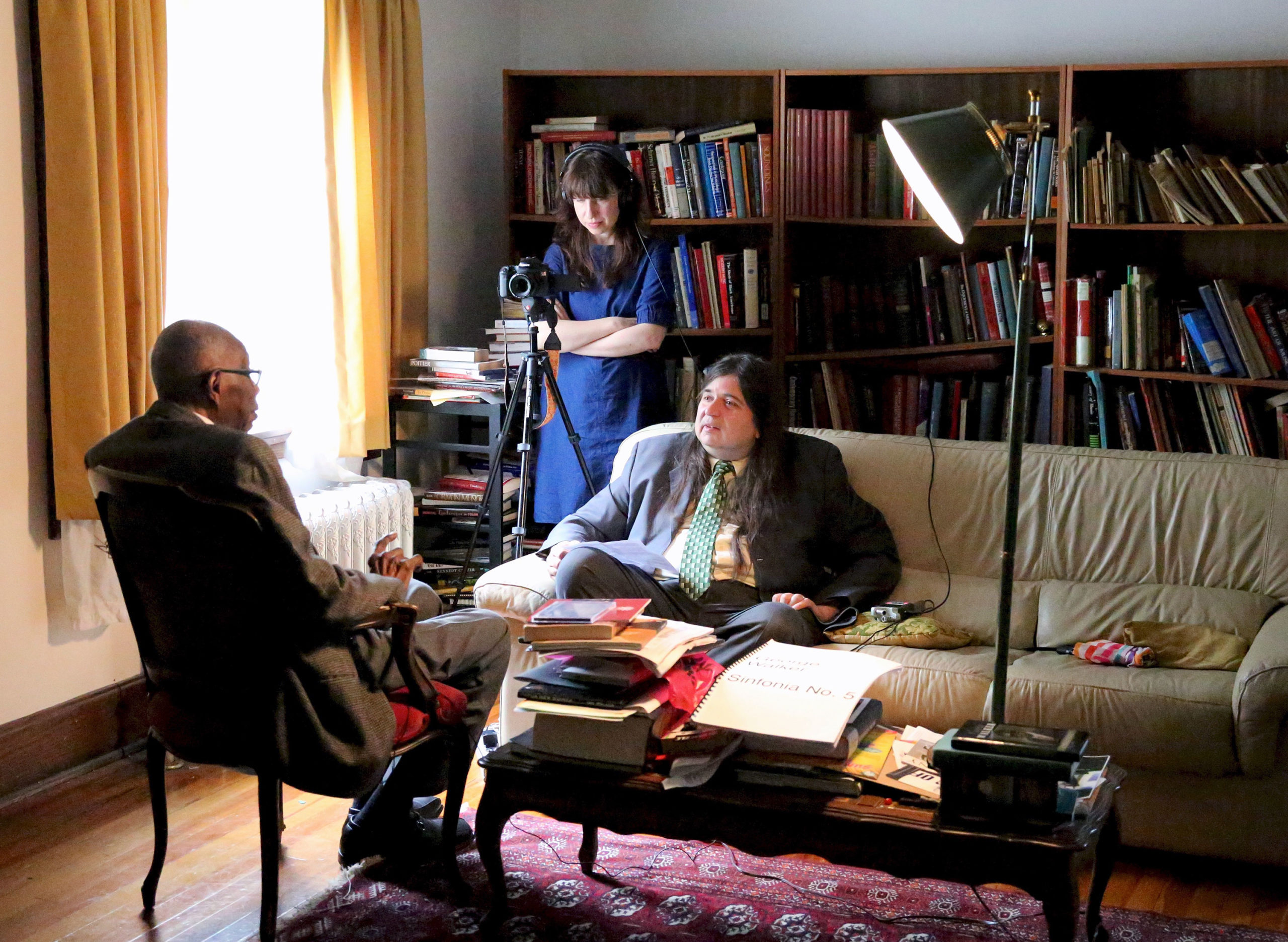 An interview takes place in a study-type room, with a man sitting on a couch, another man with his back to us sitting in a chair, and a woman in a blue dress behind the camera filming