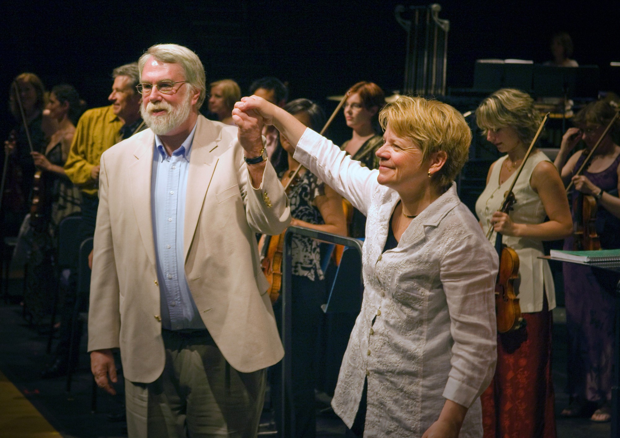 Christopher Rouse and Marin Alsop holding hands on stage in front of members of an orchestra.