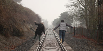 Two people photographed from the back walking on the left and right rails of traintracks in the country.