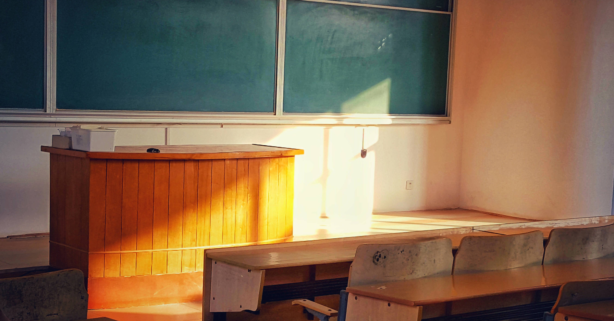 Photo by Barry Zhou (via Unsplash) of a blackboard, teacher's desk and students' desks in an empty classroom with sunlight streaming on the teacher's desk.