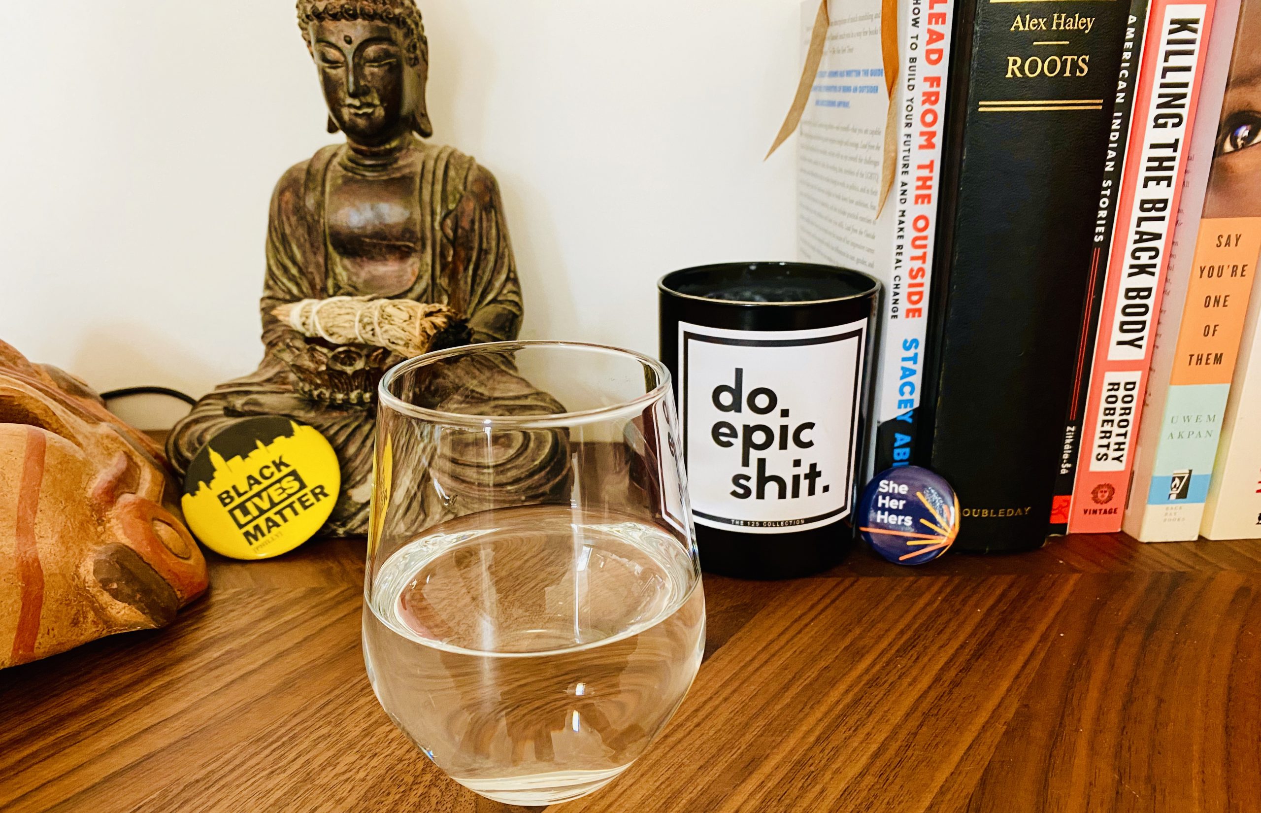 A glass of water that is half full (or empty), a mug with the caption "Do Epic Shit," a Black Lives Matter button leaning against a Buddha statue, and some books (Stavey Abrams's Lead From the Outside, Alex Haley's Roots, American Indian Stories, Dorothy Roberts's Killing the Black Body, and Uwem Akpan's Say You're One of Them), and another button ("She, Her, Hers") on a shelf in Anthony Tidd's home.