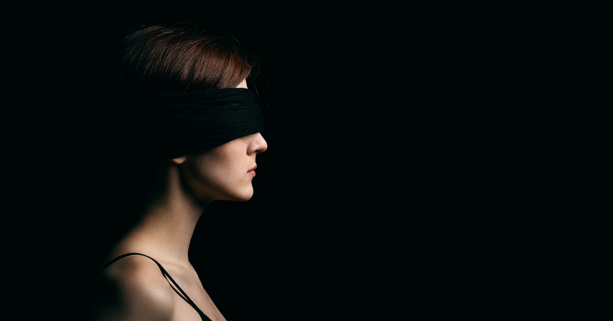 A blindfolded woman against a dark background. (Photo by Kirill Balobanov via Unsplash.)