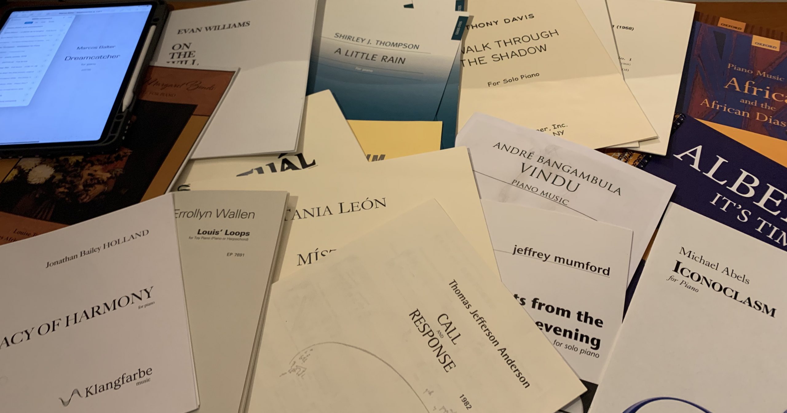 Pile of printed scores of pieces by BIPOC composers, including works by TJ Anderson, Errolyn Wallen, Tania León, Jonathan Bailey Holland, Michael Abels, and others, plus a laptop displaying a PDF score.