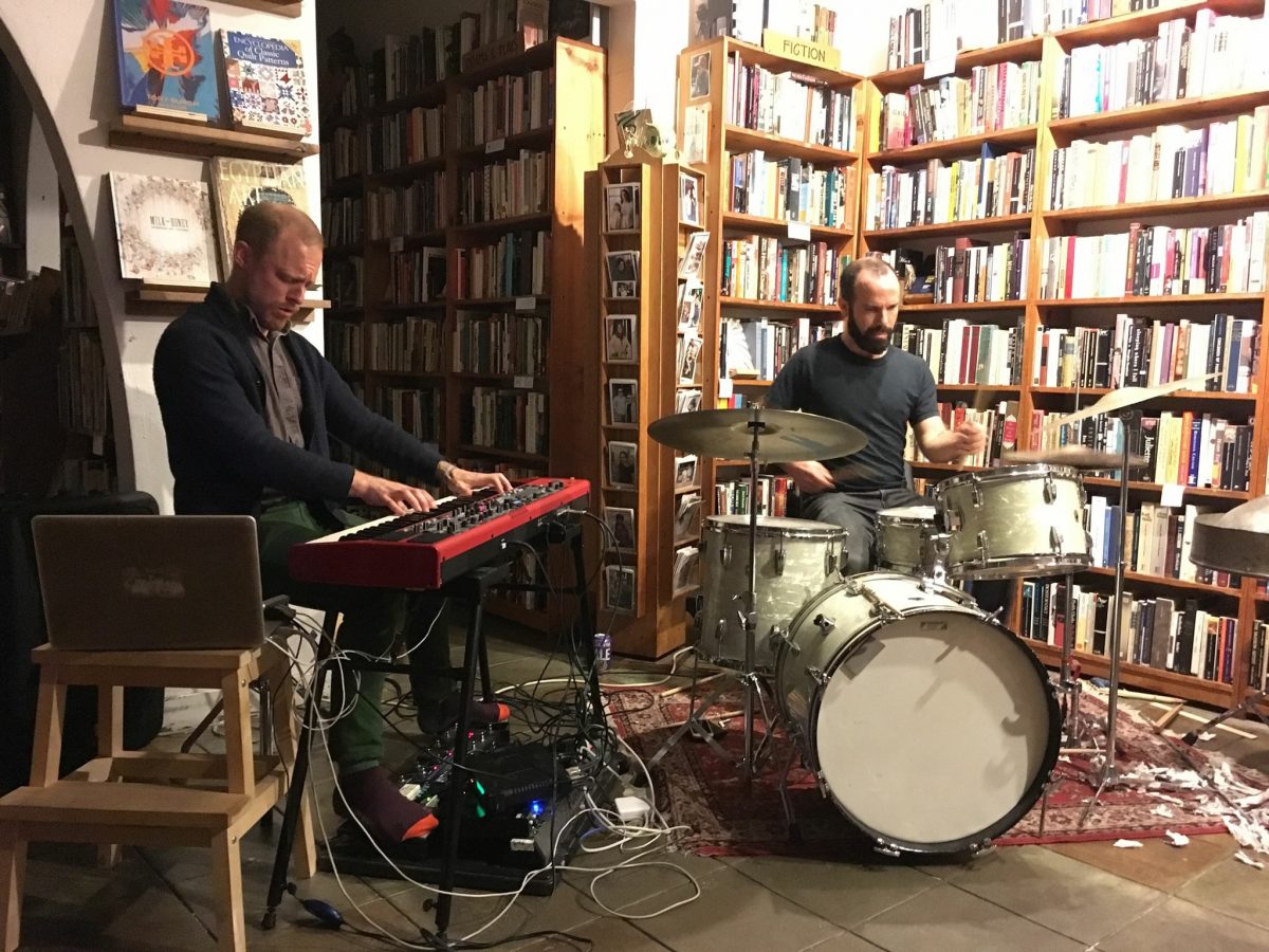 Methods Body (Luke Wyland on electric keyboard, left, and John Niekrasz on drumset, right) performing in front of rows of ceiling to floor bookshelves 