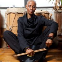 Val Jeanty holding drumsticks and sitting on a wooden floor near a window surrounded by large drums
