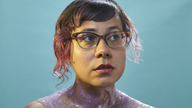 A woman with glasses and painted decoration around her neck against a teal background